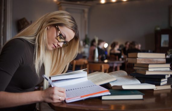 Young woman student of the University. Preparing exam and learning lessons in public library. Sitting at table surrounded by large number of textbooks