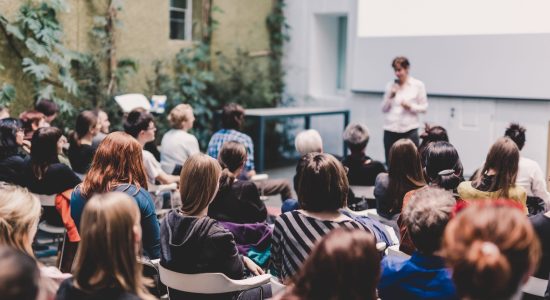 Female speaker giving presentation in lecture hall at university workshop. Audience in the conference hall. Rear view of unrecognized participant in audience. Scientific conference event.